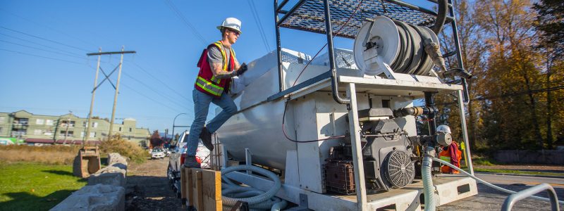 A construction worker in a safety vest and helmet operates equipment on a worksite, standing on a platform beside a large machine with hoses and control panels, under a clear blue sky.