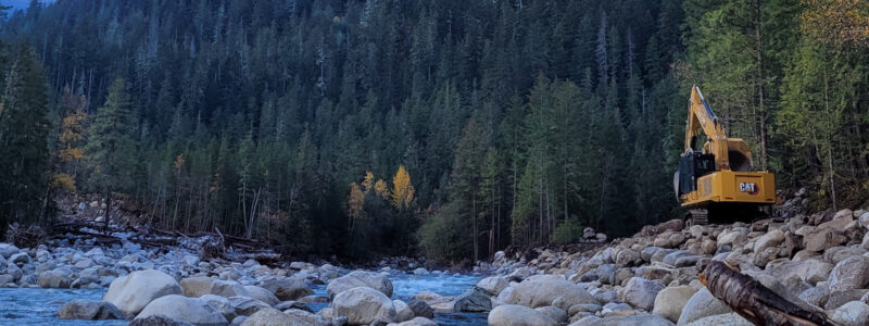 A yellow excavator is perched on large boulders beside a flowing river, surrounded by dense evergreen forest and distant mountains under a clear sky.