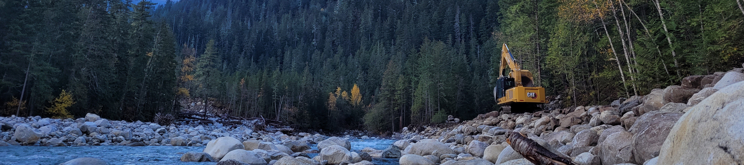 A yellow excavator is perched on large boulders beside a flowing river, surrounded by dense evergreen forest and distant mountains under a clear sky.