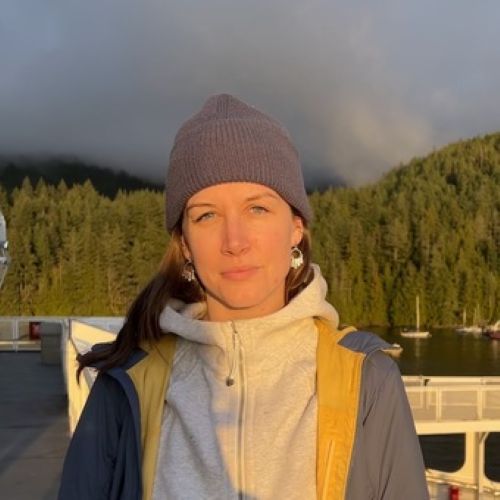 Claire Armour in a gray beanie and light jacket stands outdoors, with sunlight on her face. Behind her are green trees, water, and a misty sky, suggesting a peaceful, natural setting.