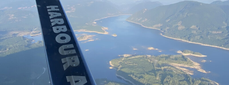 Aerial view of a winding river, forested hills, and mountains, seen through a window with the word HARBOUR visible on part of an aircraft wing or strut in the foreground.