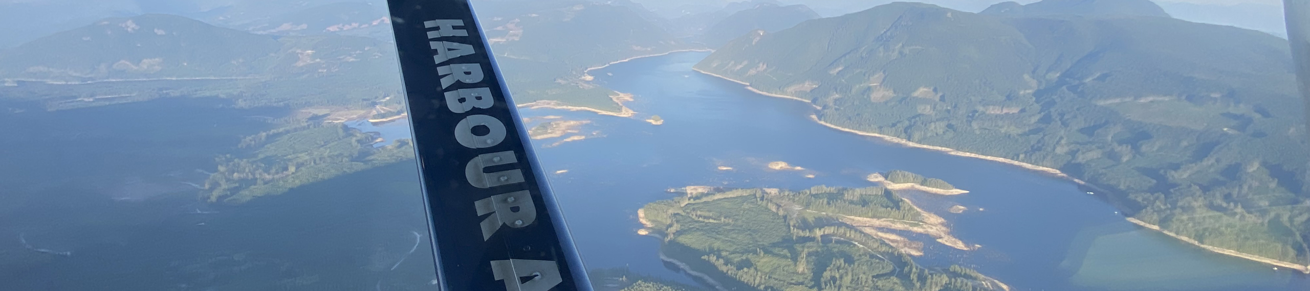 Aerial view of a winding river, forested hills, and mountains, seen through a window with the word HARBOUR visible on part of an aircraft wing or strut in the foreground.