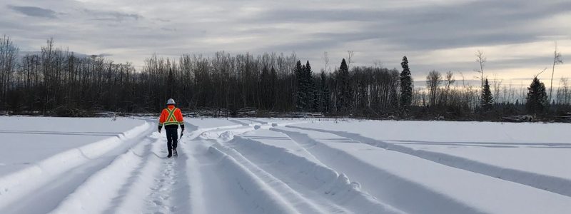 A person in a safety vest and helmet walks alone through deep snow on a wide, snowy path surrounded by trees, under a cloudy sky.