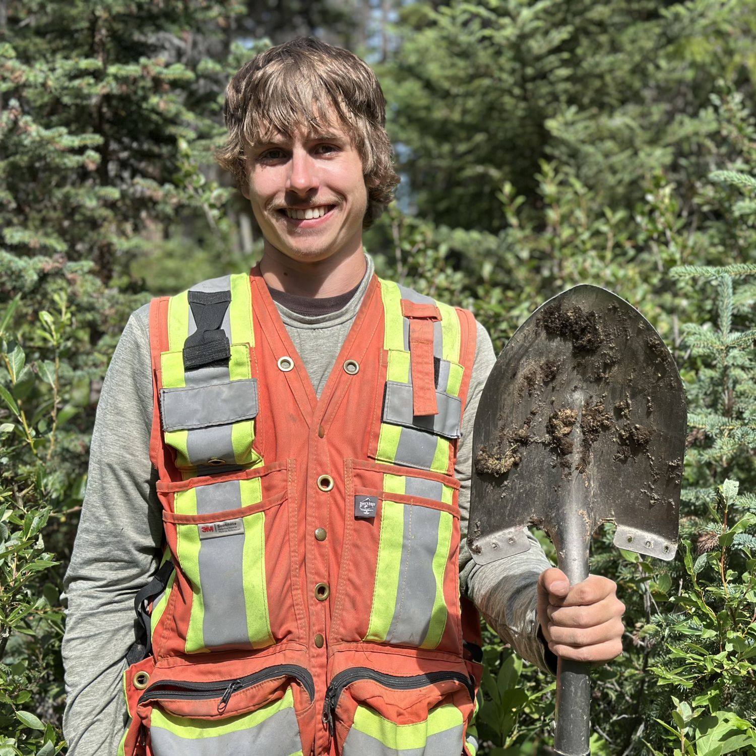 Jacob Loukianoff smiling and wearing an orange safety vest with reflective stripes stands outdoors in front of green trees, holding a shovel with dirt on its blade.