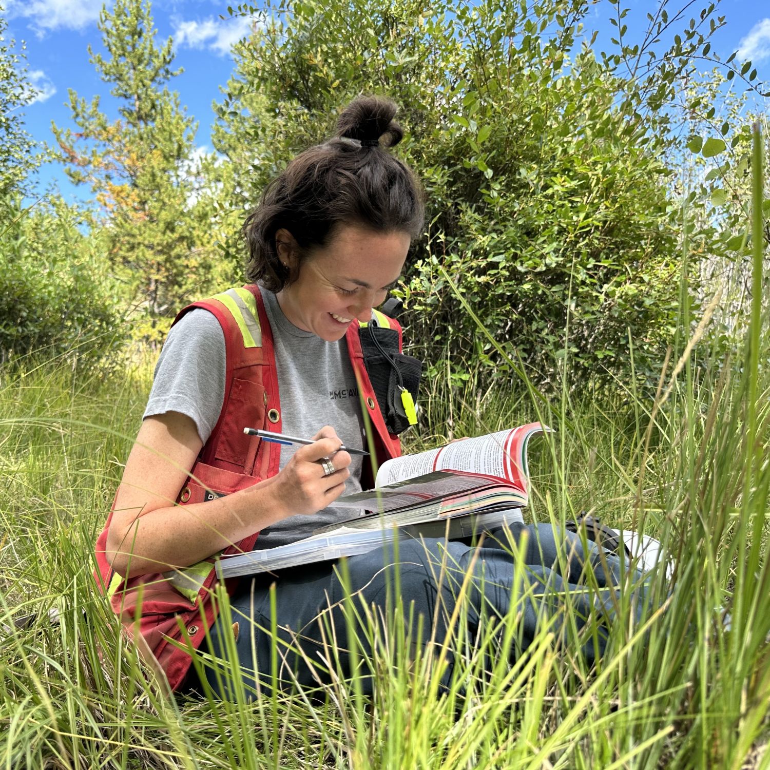 Jordyn Carss sitting down, wearing a McTavish branded T-shirt and an orange safety vest enjoying fieldwork on a sunny day.