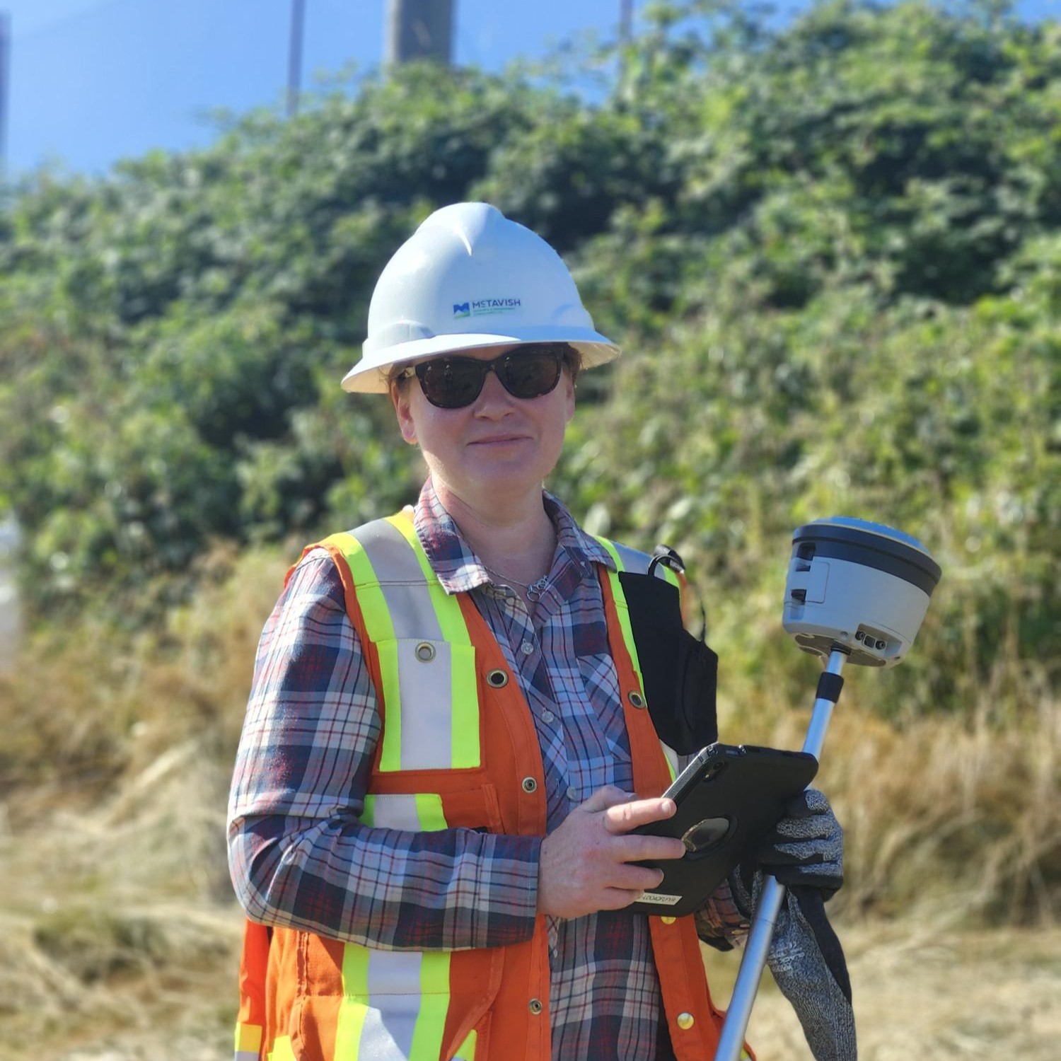 Theresa wearing a hard hat, sunglasses, and an orange safety vest holds a tablet and surveying equipment outdoors, with greenery and tall grass in the background.