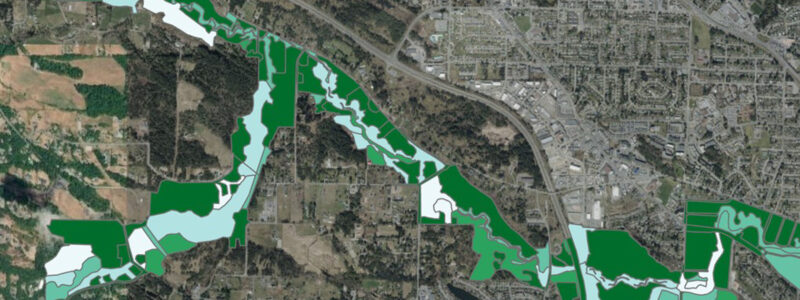 Aerial map view showing a river bordered by green and light blue highlighted areas, indicating parks, protected land, and waterways winding through a mix of urban and rural landscapes.