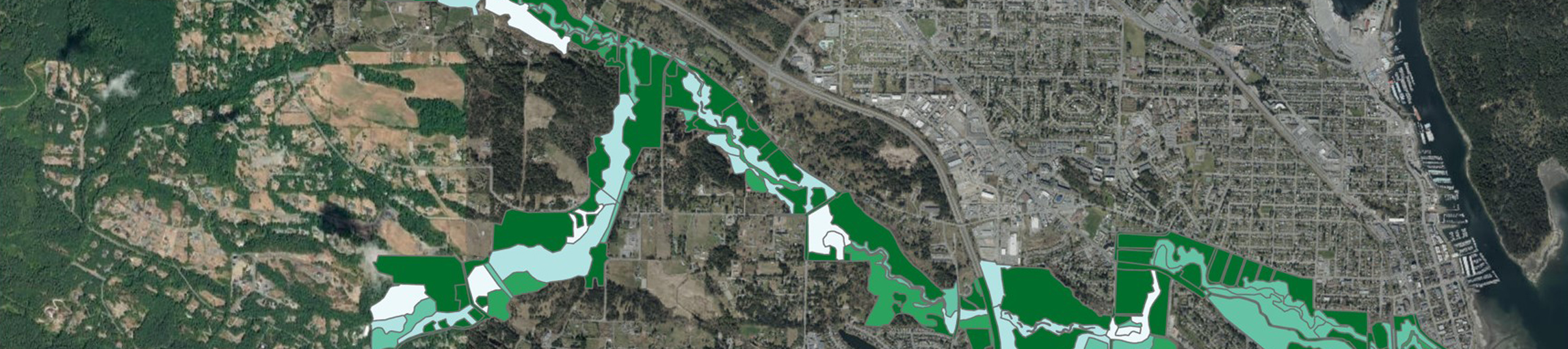 Aerial map view showing a river bordered by green and light blue highlighted areas, indicating parks, protected land, and waterways winding through a mix of urban and rural landscapes.