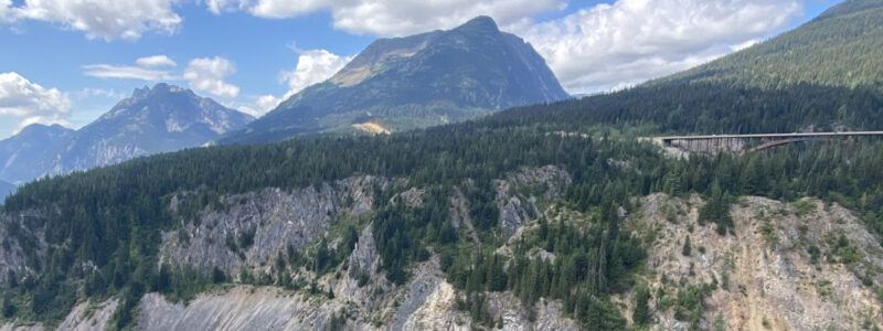 Mountain landscape with lush green forests, rocky cliffs, and a bridge crossing a deep valley; blue sky with scattered clouds and distant peaks in the background.