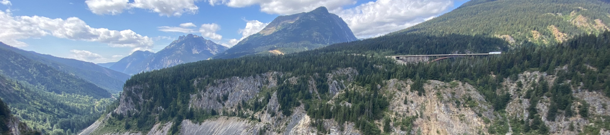Mountain landscape with lush green forests, rocky cliffs, and a bridge crossing a deep valley; blue sky with scattered clouds and distant peaks in the background.