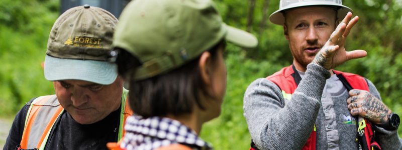 Three people in safety vests and hard hats discuss soil surveys outdoors in a green, wooded area. One gestures while speaking; another looks down, and the third listens with their back to the camera. Ideal for agriculture or arborist contexts.