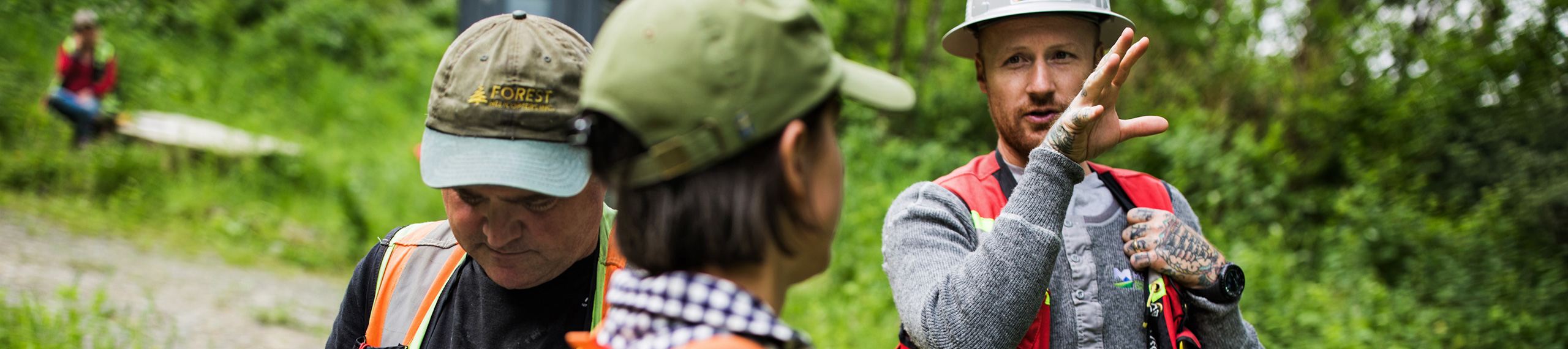 Three people in safety vests and hard hats discuss soil surveys outdoors in a green, wooded area. One gestures while speaking; another looks down, and the third listens with their back to the camera. Ideal for agriculture or arborist contexts.
