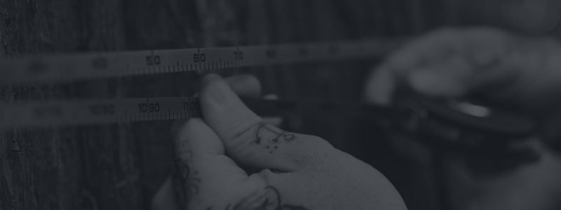 Close-up of tattooed hands holding a measuring tape against a wooden surface, with numbers visible on the tape.