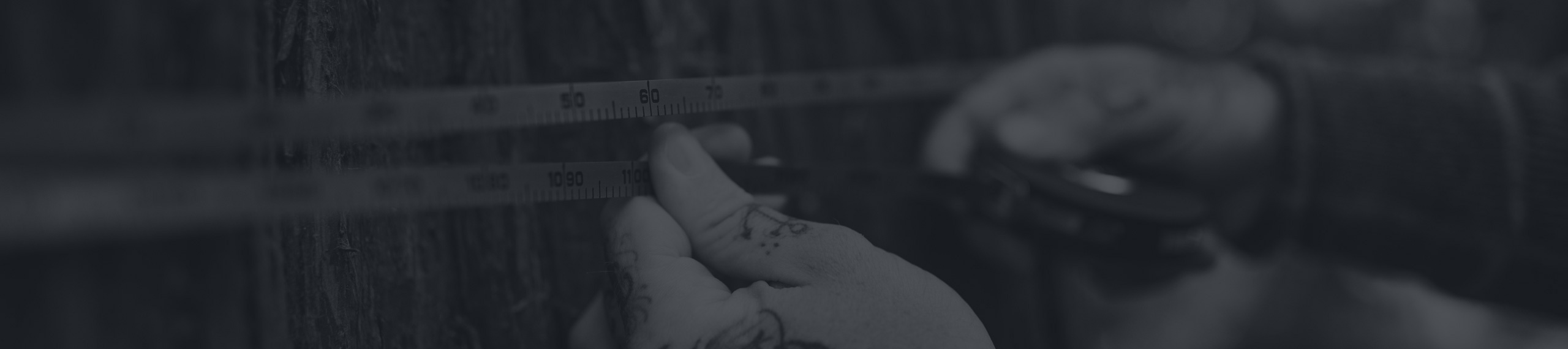 Close-up of tattooed hands holding a measuring tape against a wooden surface, with numbers visible on the tape.