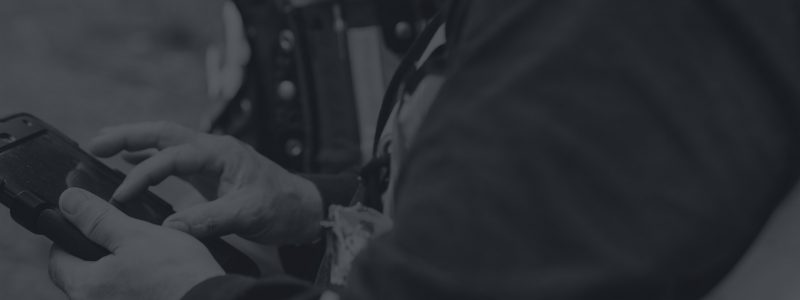 A person wearing a jacket uses a handheld device outdoors, tapping on its screen with both hands. The image is in black and white.