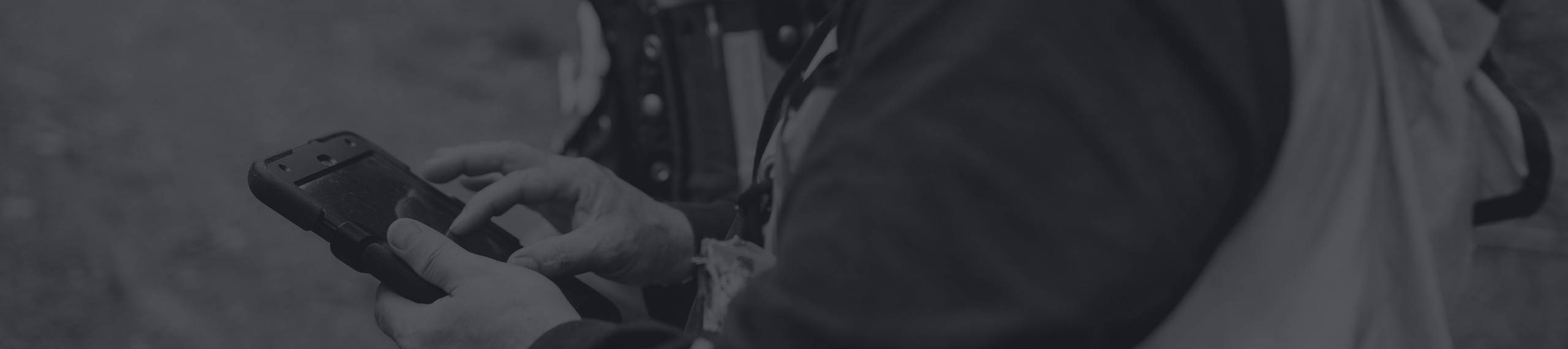 A person wearing a jacket uses a handheld device outdoors, tapping on its screen with both hands. The image is in black and white.