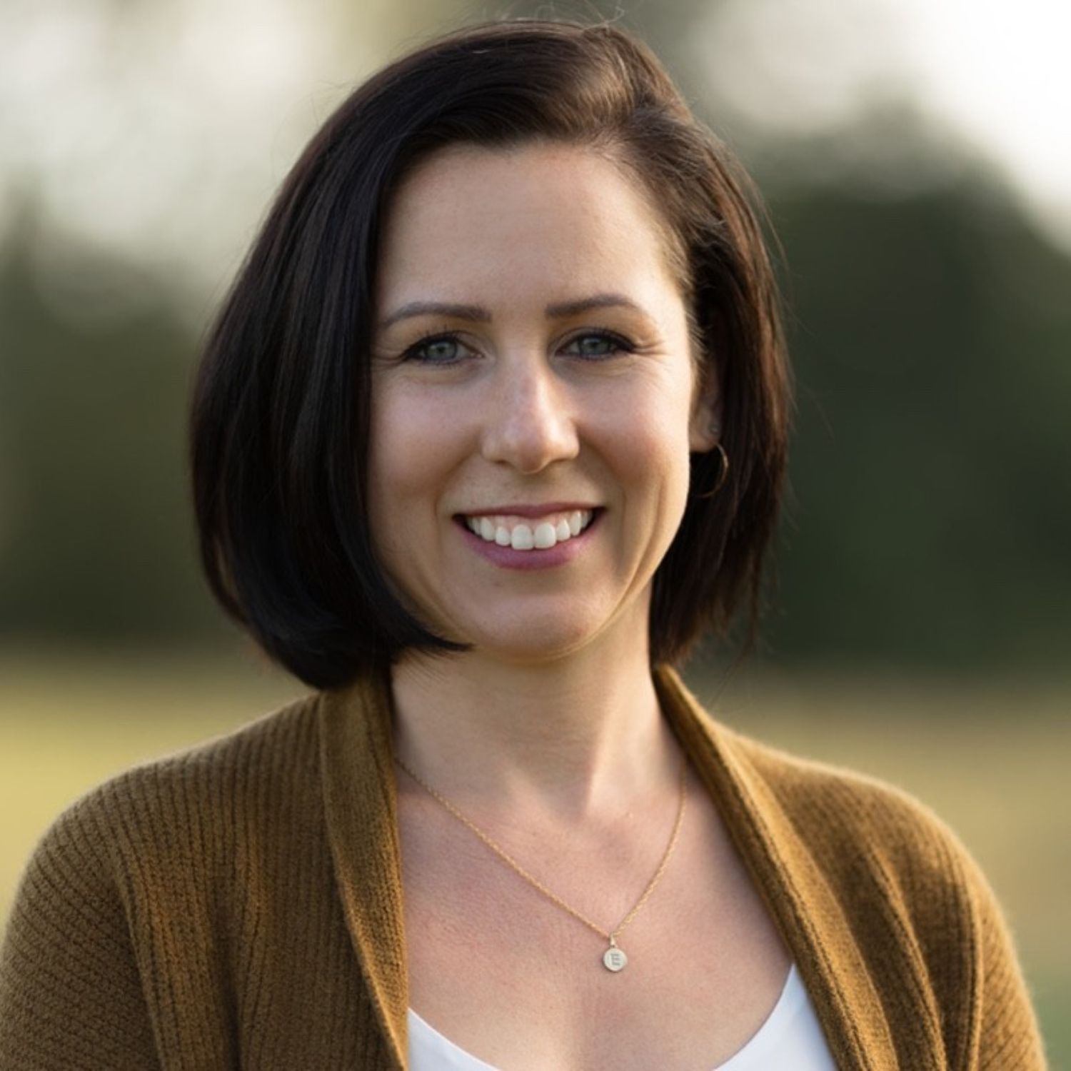 Kara Geransky smiles at the camera, wearing a brown cardigan, a white top, and a gold necklace. The background is outdoors and softly blurred with greenery.